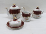 SCHMIDT- Conjunto para chá em porcelana branca esmaltada, decorado com rosáceas em policromia, detalhes em dourado com longa faixa na cor grená, composto por um bule de chá, uma leiteira, uma manteigueira, um açucareiro. Manufatura no verso. Medindo: bule para chá 15 cm alt. Mínimo bicado na borda do bule.