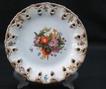 Um prato decorativo em porcelana branca, decorada ao centro com frutas, borda vazada com florais e rica douração pintadas a mão. Med. 21 cm diâmetro.