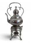 Samovar antigo em metal prateado, ricamente trabalhado com decoração em relevo e alça ornamentada. Acompanha base com suporte tripé e reservatório inferior com correntes decorativas. Peça de forte apelo decorativo, ideal para composição clássica. Mede 47 cm altura.
