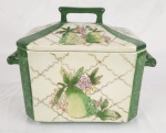 Charmosa terrina em porcelana esmaltada, com decoração policromada representando frutas e elementos florais, sobre fundo com padrão geométrico. Bordas e alças em tonalidade verde. Tampa com pegador integrado, acompanhando o desenho da peça. Medidas: 20 cm (altura) x 26 cm (comprimento) x 17 cm (largura). Apresenta sinais do tempo, com presença de fios de cabelo (craquelê) e restauro na tampa. (Colada). 