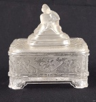 Caixa porta-joias em vidro prensado fosco, ricamente decorada em relevo floral, com tampa encimada por figura oriental estilizada. Peça de inspiração Art Déco. Apresenta restauro/colagem em uma das partes. Medidas: 18 x 17 x 11 cm. 