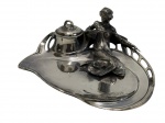 Antigo tinteiro no estilo Art Nouveau confeccionado em metal espessurado a prata, composto por base com linhas sinuosas e reservatório com tampa removível, acompanhado por figura feminina em relevo, delicado trabalho escultórico e ornamental,com acabamento polido e detalhes vazados na estrutura lateral. Medidas: 12,5 x 18 x 8 cm.