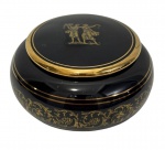 Antiga caixa em porcelana de formato circular, apresentando acabamento em tonalidade negra com delicada decoração dourada em reserva central representando figuras clássicas femininas. Medidas: 9,5 x 5 cm.