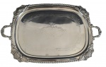 Imponente bandeja de metal espessurado a prata, de grandes dimensões, ricamente ornamentada com elegantes relevos florais e delicados arabescos gravados ao centro. Possui bordas trabalhadas em estilo clássico, com acabamento torsadé e alças laterais decoradas. Medidas: 64 x 40 x 3 cm.