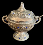 Açucareiro em metal espessurado, ricamente ornamentado com decoração floral em relevo e aplicações de pedras azuis. Corpo bojudo com alças laterais, apoiado sobre base elevada, acompanhado de tampa e colher. Med.: 13,5 x 9,5 x 12,5 cm E Colher: 13 cm.