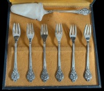 Conjunto para sobremesa composto por seis garfinhos e uma pá para bolo em metal espessurado a prata, acondicionado em estojo original revestido internamente em veludo. As peças apresentam elegante decoração em relevo de inspiração rococó nos cabos.