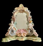 Antigo espelho de mesa em faiança art nouveau, ricamente ornamentado com volutas em relevo, policromia em tons suaves e aplicações figurativas de querubins laterais, apresentando espelho central recortado e decoração floral no coroamento. Peça de grande efeito decorativo, com marcas do tempo. Medidas: 48 x 44 x 16,5 cm. Apresenta bicado e craquelado.