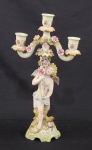 Castiçal decorativo em biscuit policromado, composto por figura de querubim com flores em relevo sustentando estrutura superior com três braços porta-velas em tom verde claro e detalhes dourados, medindo 44 cm de altura e 25 cm de largura. Discretos bicados.