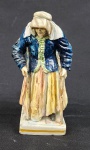 Estatueta napolitana Capodimonte, representando Mendiga vestida com roupas esfarrapadas, um adorno de cabeça tradicional e duas bengalas, feita de porcelana esmaltada e policromada. Medindo 17 cm de altura, com avaria na bengala direita, perda de esmalte e craqueamento.