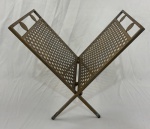 Revisteiro articulado em metal com acabamento em pátina ouro-velho. Estrutura em `X` (estilo tesoura), permitindo o fechamento para armazenamento. Laterais decoradas com trama em treliça vazada (padrão romboide). Alças superiores integradas à estrutura co