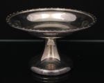 Fruteira de pé alto confeccionada em metal espessurado à prata, centro ligeiramente côncavo, borda relevada com flores intercaladas por perolados, fuste afunilado sobre base circular alteada. Ausência de punção ou marca de manufatura. Medidas: Altura: 12 x Diâmetro 21 cm.