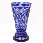 Vaso executado em cristal overlay, double de azul cobalto e incolor; corpo obcônico sobre base circular alargada, fundo preenchido com estrela de doze pontas; decoração em lapidação composta por elementos geométricos e radiais, incluindo painéis facetados, motivos em leque, losangos e reservas ovais na borda e no pé. Interior incolor visível nas áreas lapidadas. Apresenta desgaste aparente na área da base, vide foto. Medidas: Altura: 29 x Diâmetro: 16 cm.