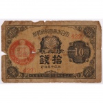 Cédula japonesa  10 Sen, emissão do período imperial (início do século XX, provável era Taish / início Shwa). Anverso: composição com moldura ornamental, numerais de valor nos cantos e cartela lateral. Ao centro, representação do crisântemo imperial (Kikumon), emblema da Casa Imperial Japonesa, inserido em arranjo vegetal estilizado. Campo esquerdo com selo vermelho oficial (carimbo de validação/controle).  Inscrições em caracteres japoneses verticais indicando valor e autoridade emissora. Numeração impressa em vermelho. No estado, apresenta rasgos, vincos, e perda da legibilidade da impressão do reverso. Medidas: Altura: 5,3 x Comprimento: 8,5 cm.