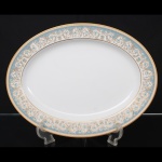 NORITAKE, Nagoya - Japão(Fundada em 1904 pelos irmãos Ichizaemon e Toyo Morimura). Exuberante travessa japonesa em porcelana da década de 1960/70, padrão POLONAISE (2045); Formato oval, raso, de centro ligeiramente rebaixado. Aba profusamente ornamentada por composição vegetalista, composta por rinceau, volutas, ramos, folhas e flores dispostas em repetição, sobre cartela de tonalidade azul ciano, seguindo repertório derivado de padrões europeus, estilo neoclássico e revivalista; contornos e elementos internos definidos por generosa aplicação de filetes à ouro, incluindo a borda e o centro. Marca da manufatura no verso. Medidas: Comprimento: 29,5 x Largura: 22,5 cm.