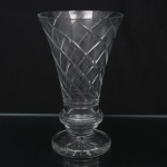 Elegante vaso executado em meio cristal, diáfano e incolor; bojo obcônico com abertura larga, talhado com sulcos e frisos em espiral, fuste anelar facetado com janelas oblongas, base circular espessada, fundo preenchido com estrela de dezesseis pontas. Apresenta três acentuados bicados na borda, demanda restauro. Estado conforme fotografias. Medidas: Altura: 23 x Diâmetro: 14 cm.