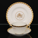 Minton, Staffordshire - Inglaterra. Excepcional trio para chá em porcelana inglesa da década de 1950, padrão Gold Rose. Esmaltagem vítrea de tom branco perolado, realçado por profusa ornamentação com faixas e filetes à ouro, fundo interno contendo flor em reserva. Bojo aproximadamente obcônico com alça lateral em voluta, assente sobre base circular, ricamente modelado com frisos em espiral relevados na própria pasta; padrão decorativo replicado no pires. Marca da manufatura no verso. Medidas: Xícara: Altura: 5,5 x Comprimento: 11 x Largura: 9 cm; Pires: Diâmetro: 14 cm; Prato: Diâmetro: 17,5 cm.(apresenta dois raso trincado na base do bojo, e leves manchas do tempo).