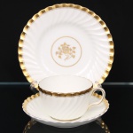 Minton, Staffordshire - Inglaterra. Excepcional trio para chá em porcelana inglesa da década de 1950, padrão Gold Rose. Esmaltagem vítrea de tom branco perolado, realçado por profusa ornamentação com faixas e filetes à ouro, fundo interno contendo flor em reserva. Bojo aproximadamente obcônico com alça lateral em voluta, assente sobre base circular, ricamente modelado com frisos em espiral relevados na própria pasta; padrão decorativo replicado no pires. Marca da manufatura no verso. Medidas: Xícara: Altura: 5,5 x Comprimento: 11 x Largura: 9 cm; Pires: Diâmetro: 14 cm; Prato: Diâmetro: 17,5 cm.