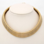 Gargantilha articulada em metal dourado, composta por sucessão de gomos tubulares canelados, dispostos de maneira contígua em conformação semicircular flexível. Estrutura de perfil escalonado, com acabamento polido e fecho integrado por sistema de encaixe. Medida: Diam.: 12,5 cm.