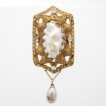 Broche em metal dourado de composição vazada, estruturado em moldura retangular recortada por motivos fitomorfos e volutas estilizadas, centrada por madrepérola ovalada de contorno irregular, fixada por garras recortadas. Campo ornado por aplicação de pedras incolores facetadas. Arremate inferior pendente em formato periforme perolado, suspenso por correntes de elos miúdos. Reverso com fecho de alfinete horizontal. Medidas: Comp.: 9,5 x Larg.: 4,5 cm.
