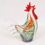 Escultura animalista representando galo, executada em vidro artístico de Murano, soprado e moldado, corpo translúcido com aplicações policromadas em tons de verde, azul, laranja e vermelho, apresentando decoração interna e penas estilizadas. Crista e barbela em vidro vermelho translúcido, cauda recurvada incolor. Apresenta dois discretos trincados no fundo da base. Medidas: Alt.: 24 x Comp.: 21 x Larg.: 8 cm.