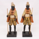 Par de esculturas executadas em material sintético policromado, representando pajens mouros portando bastões, trajando turbantes, capas, gibões e calções listrados de feição seiscentista. Figuras masculinas apresentadas em posição de marcha, sobre bases retangulares escalonadas. Policromia em tons preto, ocre, dourado e marfim. Medidas: Alt.: 37 x Comp.: 10,5 x Larg.: 15cm.