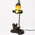 Luminária de mesa em composição de estilo Art Nouveau, estruturada em metal patinado, com haste sinuosa sustentando tulipa em vidro moldado e acidado, com policromia em tonalidades verde e âmbar, ornamentada por motivos fitomórficos em relevo. Base recortada apresentando figura de putto alado em posição reclinada. Parte elétrica funcionando. Medidas: Alt.: 43 x Comp.: 20 x Larg.: 20cm.