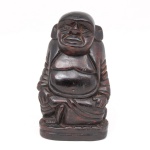 Escultura executada em bloco de madeira entalhada representando Budai (Hotei), figura associada ao budismo zen e popularmente identificada como Buda da felicidade, representado sentado. Possui desgastes e marcas do tempo. Medidas: Alt.: 20 x Comp.: 12 x Larg.: 9 cm.