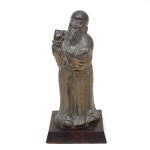 Escultura oriental em metal patinado representando Shou Lao, divindade chinesa associada à longevidade, trajando vestes ornamentadas e portando pêssego simbólico. Figura com longa barba, apoiada sobre base retangular em madeira. Apresenta marcas de uso. Medidas: Alt.: 22 x Comp.: 10 x Larg.: 10 cm.