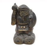 Escultura executada em metal patinado representando Daikoku, uma das Sete Divindades da Fortuna da tradição japonesa, associado à prosperidade e abundância. Figura trajando vestes tradicionais, portando malho da fortuna (uchide no kozuchi) e saco às costas, apoiada sobre fardos de arroz, símbolo de riqueza e fertilidade. Medidas: Alt.: 13,5 x Comp.: 8,5 x Larg.: 7,5 cm.