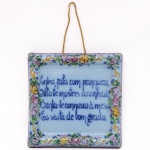 Placa de parede em faiança portuguesa da região de Alcobaça, de formato quadrangular, com decoração policroma em esmaltação manual, apresentando cercadura floral e inscrição central em azul cobalto: Entra, fala com franqueza, não te mostres acanhado, senta-te connosco à mesa, és visita de bom grado. Verso identificado Alcobaça, Made in Portugal, numerado 286. Apresenta cordão para suspensão em parede. Medidas: Alt.: 17,5 x Comp.: 18 cm.