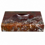 Cinzeiro de mesa em pedra polida, possivelmente jaspe, de formato retangular, com cavidade central oval escavada. Corpo monolítico com inclusões minerais e manchas naturais em tonalidades avermelhadas, ocres, verdes, negras e esbranquiçadas. Superfície lapidada e polida, com laterais retas. Peça destinada ao acondicionamento de cinzas e apoio para cigarro. Medidas: Alt.: 3,5 x Comp.: 22,5 x Larg.: 16 cm.
