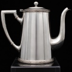 WOLFF. Elegante bule para café confeccionados em metal espessurado à prata, bojo aproximadamente cônico, bordas contornadas por barrados relevados com frisos intercalados por flores, padrão conhecido pelo senso comum como "florzinha"; tampa dotada de abertura através de movimento basculante, puxador circular, bico curvilíneo de ponta recortada, alça lateral curvada com descanso de polegar retilíneo. Marca da manufatura puncionada ao fundo. Meds.: Alt. 18,5 x Comp. 22  x Larg. 10,5 cm.