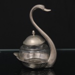 Pote para geléia com suporte em metal prateado, executado na forma de cisne, recipiente removível em vidro moldado de corpo bojudo, tampa também em metal. Medidas: Alt.: 16 x Comp. 12 x Larg. 8,5 cm. Recipiente: Alt.: 9,5 x Diam.: 7,5 cm.