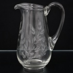 CRISTAIS HERING. Elegante jarra da década de 1960/70 executada em meio cristal, diáfano e incolor; bojo balaústre talhado com padrão de folhagens estilizadas, borda ondulada, bico vertedor revirado para fora, alça lateral curvada aplicada sobre a superfície. Medidas: Alt.: 23 x Comp.: 18 x Larg.: 13 cm.