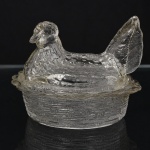 Manteigueira em vidro prensado incolor, composto por base ovalada simulando cesta entrançada e tampa modelada na forma de galinha pousada, com representação naturalista da plumagem, cabeça e cauda em relevo. Medidas: Alt.: 11 x Comp.: 14 x Larg.: 11 cm.
