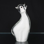 Escultura em vidro artístico soprado e moldado, representando urso polar em composição estilizada. Corpo em vidro translúcido incolor envolvendo núcleo interno em vidro branco leitoso, executado em técnica de sommerso. Cabeça elevada com focinho proeminente, orelhas laterais aplicadas e olhos em vidro preto. Modelagem de contornos curvilíneos e volumes compactos, assente sobre base integrada ao corpo. Medidas: Alt.: 24,5 x Comp.: 13,5 x Larg.: 7 cm.