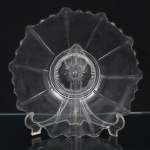 Prato de serviço em vidro incolor prensado, de formato circular e perfil raso, apresentando borda ondulada recortada em movimentos sucessivos. Corpo ornamentado por gomos radiais em relevo que convergem para medalhão central decorado com motivo floral estilizado, circundado por faixa canelada. Medida: Diam.: 31 cm.