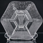 Petisqueira em vidro incolor prensado, de formato hexagonal, compartimentada em seis divisões periféricas dispostas ao redor de reservatório central igualmente hexagonal. Corpo decorado em relevo com padrão contínuo de folhas nervuradas e pequenas flores estilizadas distribuídas por toda a superfície externa. Produzida por prensagem em molde, apresenta desenho geométrico de inspiração naturalista, combinando organização radial e ornamentação vegetal. Destinada ao serviço de aperitivos, confeitos, frutas secas ou pequenas porções, permitindo a disposição simultânea de diferentes alimentos. Medidas: Diam.: 31 cm.