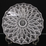 Prato decorativo em cristal incolor prensado, de formato circular com borda recortada em múltiplos segmentos angulados, ornamentado por composição radial em relevo, formada por sucessivas reservas ogivais e losangulares que abrigam elementos esféricos salientes, distribuídos concentricamente ao redor de medalhão central circular. Medida: Diam.: 29,5 cm.