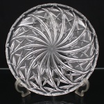 Prato circular em cristal incolor prensado, com borda recortada e decoração de composição radial; campo central formado por conjunto de caneluras curvas dispostas em movimento espiralado convergente, criando efeito de roseta dinâmica, ao redor, distribuem-se motivos estrelados de quatro pontas em repetição regular. Medida: Diam.: 29 cm.