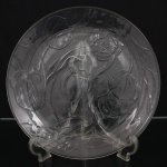 Prato circular em vidro prensado translúcido, decorado em relevo com composição floral de inspiração Art Nouveau, apresenta flores de tulipa em diferentes estágios de abertura, acompanhadas por hastes sinuosas e folhagem estilizada que se distribuem por toda a superfície, formando desenho contínuo integrado ao campo e à aba. Medida: Diam.: 33 cm.