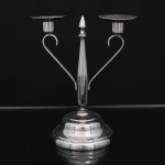 Castiçal para duas velas em metal prateado, formato geométrico e composição simétrica, base circular escalonada, haste central facetada encimada por pináculo cônico e dois braços arqueados em disposição aberta, terminados em bocais cilíndricos com pratos coletores circulares. Medidas: Alt.: 20 x Comp.: 17 x Larg.: 10 cm.