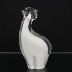 Escultura em vidro artístico soprado e moldado, representando urso polar em composição estilizada. Corpo em vidro translúcido incolor envolvendo núcleo interno em vidro branco leitoso, executado em técnica de sommerso. Cabeça elevada com focinho proeminente, orelhas laterais aplicadas e olhos em vidro preto. Modelagem de contornos curvilíneos e volumes compactos, assente sobre base integrada ao corpo. Medidas: Alt.: 24,5 x Comp.: 13,5 x Larg.: 7 cm.