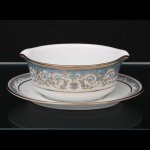 NORITAKE, Nagoya - Japão(Fundada em 1904 pelos irmãos Ichizaemon e Toyo Morimura). Exuberante molheira em porcelana da década de 1960/70, padrão POLONAISE (2045); formato oblongo assente sobre presentoir oval, vertedouros opostos revirados para fora; esmaltagem vítrea na cor branca, profusamente ornamentada por composição vegetalista, composta por rinceau, volutas, ramos, folhas e flores dispostas em repetição, sobre cartela de tonalidade azul turquesa, realçados por rica aplicação de douração, seguindo repertório derivado de padrões europeus, estilo neoclássico e revivalista; bordas contornadas com generosa aplicação de filetes à ouro e faixa perolada pontilhada, padrão decorativo integralmente replicado no presentoir. Marca da manufatura no verso. Medidas:  Alt.: 214 x Comp.: 21,5  x Larg.: 16 cm.