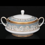 NORITAKE, Nagoya - Japão(Fundada em 1904 pelos irmãos Ichizaemon e Toyo Morimura). Exuberante terrina com tampa em porcelana da década de 1960/70, padrão POLONAISE (2045); formato aproximadamente campaniforme assente sobre base circular alteada, tampa ligeiramente abaulada encimada por puxador obcônico, alças laterais curvadas; esmaltagem vítrea na cor branca, profusamente ornamentada por composição vegetalista, composta por rinceau, volutas, ramos, folhas e flores dispostas em repetição, sobre cartela de tonalidade azul turquesa, realçados por rica aplicação de douração, seguindo repertório derivado de padrões europeus, estilo neoclássico e revivalista; bordas contornadas com generosa aplicação de filetes à ouro e faixa perolada pontilhada. Marca da manufatura no verso. Medidas:  Alt.: 14  x Comp.: 26 x Larg.: 21 cm.