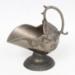 Açucareiro para torrões em metal prateado repuxado e cinzelado, de corpo ovalado sobre pedestal circular, apresentando bordas recortadas e alça móvel arqueada. Ornamentação em relevo com rosas, folhagens e volutas ao estilo do período Art Nouveau, acompanhada por friso perolado e cercadura com motivos geométricos estilizados na base. Medidas: Alt.: 15 x Comp.: 15 x Larg.: 9 cm.