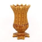 Vaso em faiança vitrificada de tonalidade ocre, corpo campaniforme com decoração em relevo composta por elementos estilizados verticais, borda recortada em ondulações e base moldada. Marca em relevo na pasta. Medidas: Alt.: 29 x Diam.: 17,5 cm.
