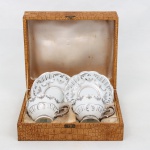 Porcelana Schmidt - Brasil(Fundada em 1945). Par de xícaras para café em porcelana nacional, esmaltação branca, decoração com reservas e ornatos prateados, asas moldadas e inscrição comemorativa Bodas de Prata no interior das xícaras. Acompanhadas de estojo original revestido em material sintético imitando couro de crocodilo. Marca da manufatura no verso. Medidas: Xícara: Alt.: 4,5 x Comp.: 8,5 x Larg.: 7 cm. ; Pires: Diam.: 10,5 cm.