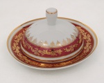 Porcelana Schmidt - Brasil(Fundada em 1945). Manteigueira em porcelana nacional da década de 1960/70; esmaltagem vítrea na cor branca, decorado com faixas de tom rubi e ornamentação em douradura aplicada, formando cercaduras de arabescos, elementos fitomorfos e rendilhados estilizados; bordas contornadas por filetes à ouro, formato abaulado encimada por pega cilíndrica, presentoir circular. Marca da manufatura no verso. Medidas: Alt.: x Diam.: