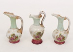 Três pequenas jarras em porcelana esmaltada e iridescente, de formato bojudo com gargalo alongado e bocal recortado, decoradas com reservas figurativas galantes e ornatos dourados. Bases circulares em tom vinho e alças modeladas. Medidas: Alt.: 8,5 x Comp.: 4 cm.